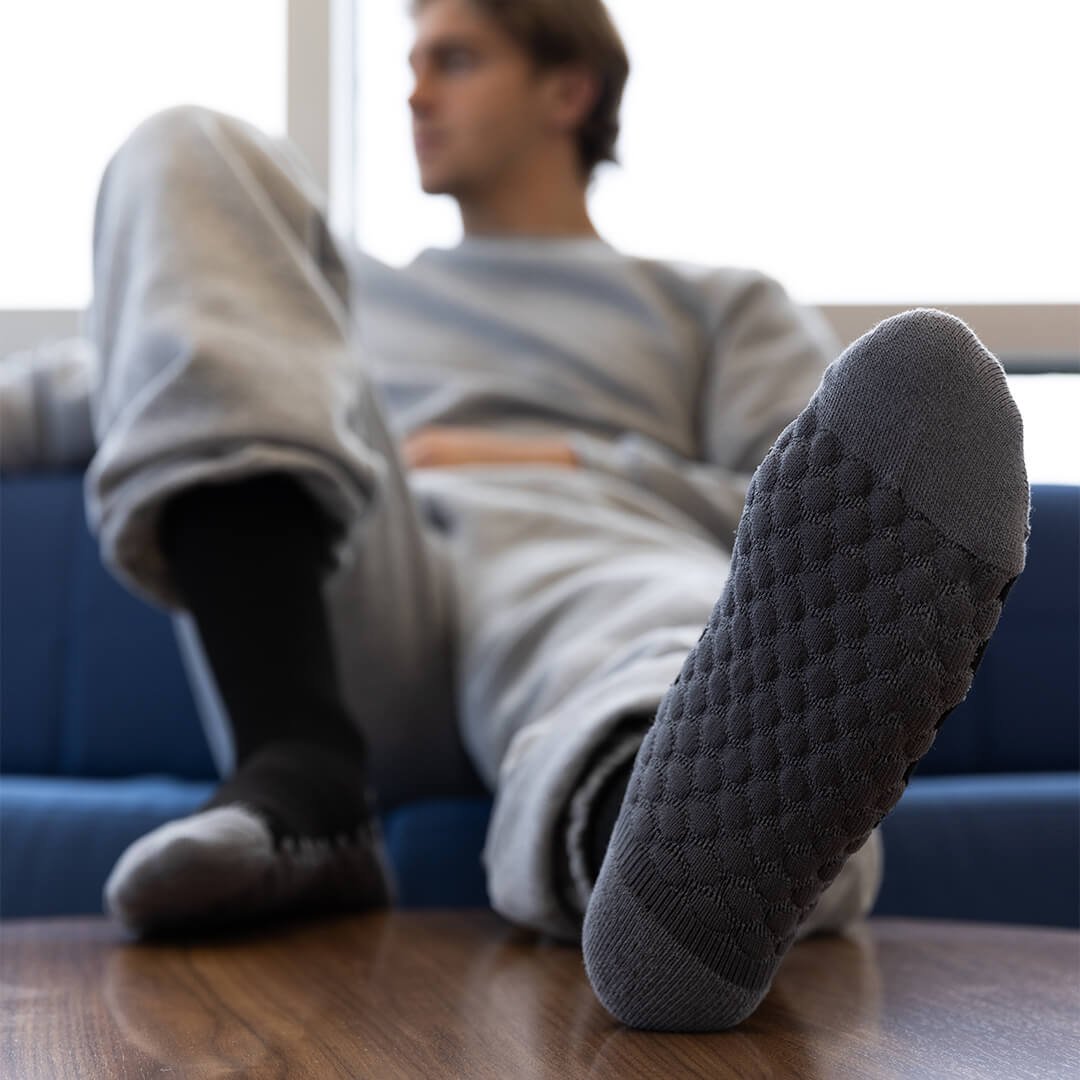 A man reclining on a blue couch with one foot raised, wearing dark gray textured knit socks. The foreground foot is sharply focused, showing the textured sole and toe, while the rest of the scene is softly blurred.