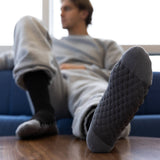 A man reclining on a blue couch with one foot raised, wearing dark gray textured knit socks. The foreground foot is sharply focused, showing the textured sole and toe, while the rest of the scene is softly blurred.