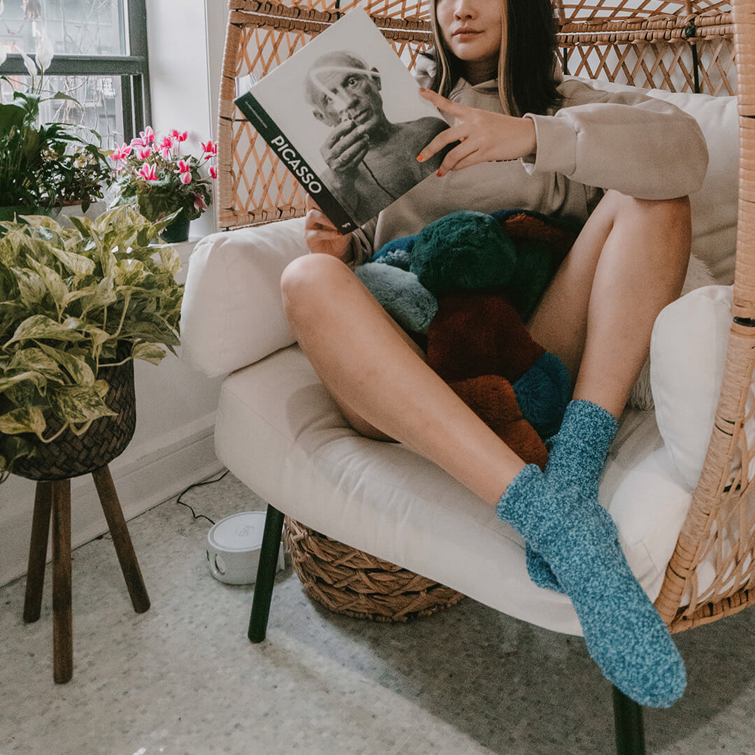 Person sitting on a white chair wearing fuzzy socks reading a book with plants and a wicker chair in the background.