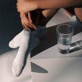 A person sitting on white geometric steps wearing white crew socks, with a clear textured glass of water beside them. The brand name 'Dr. Scholl's' is visible on the toes of the socks.