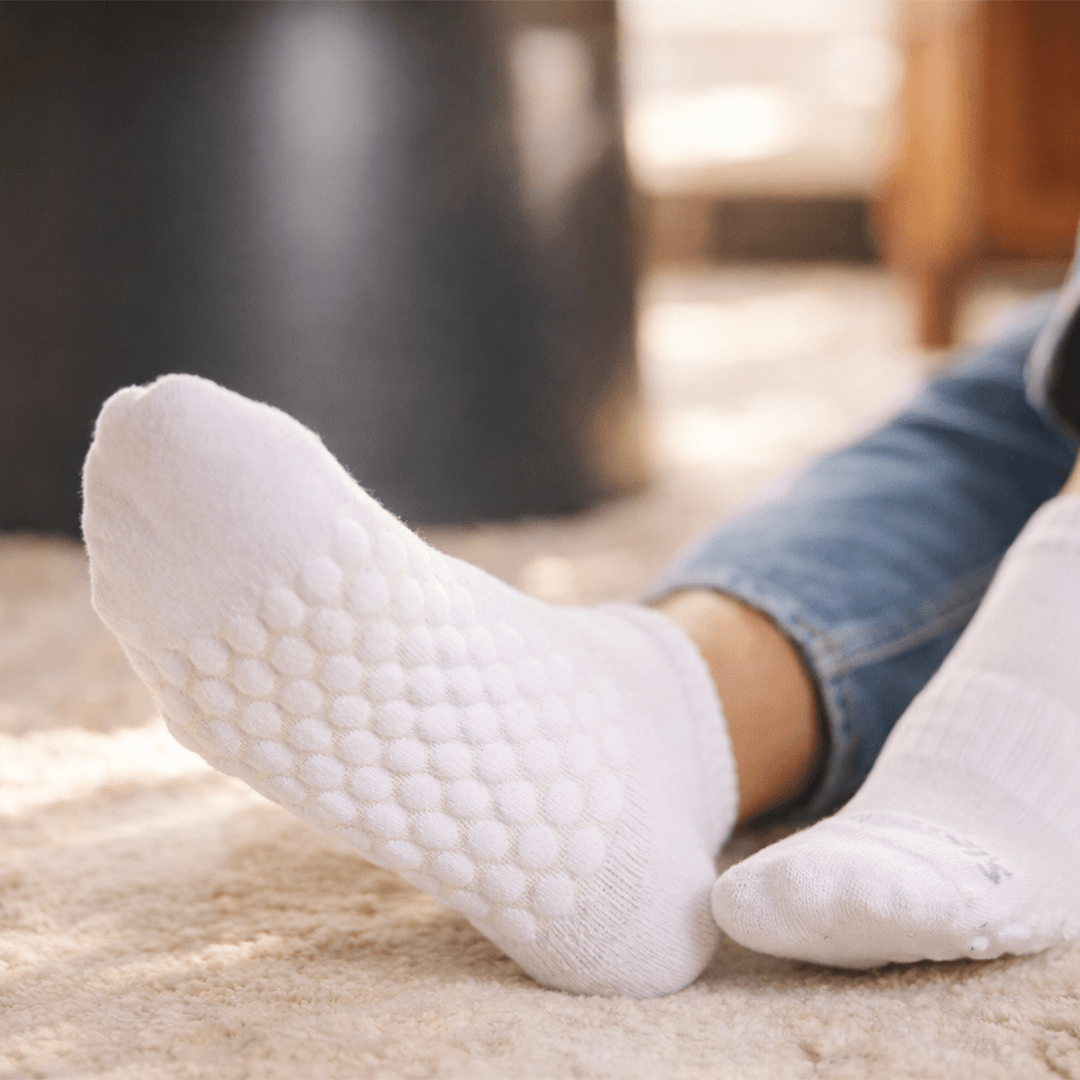 White textured grip socks worn on a person's feet, shown in a cozy indoor setting with blue jeans and a soft beige rug.