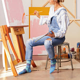 A woman sitting on a metal stool in an art studio, wearing blue denim overalls and striped blue-and-white long-sleeve shirt, holding paintbrushes. She is positioned next to a wooden easel with pink and orange paint on the canvas, with other art supplies on a stool behind her.