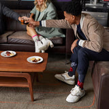 Three people gathered in a lounge setting, with one person reaching across a wooden coffee table to toast with a glass of beer. The person wears a beige jacket, navy pants, white sneakers, and red socks with white socks.