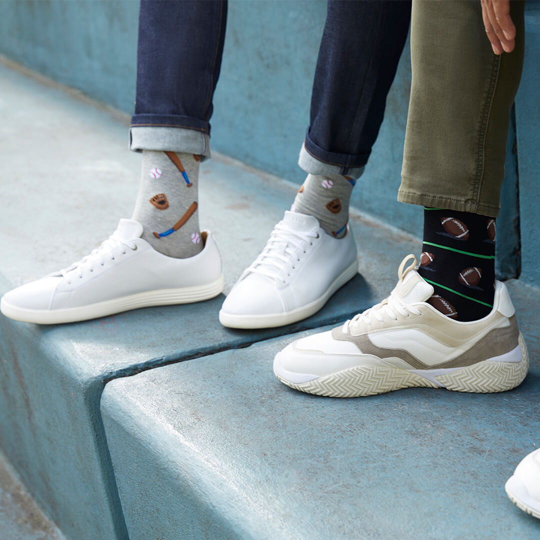 Two people standing on blue concrete steps, wearing patterned crew socks and sneakers. One person wears gray socks with baseball motifs and white sneakers, while the other wears black socks with football motifs and cream sneakers.