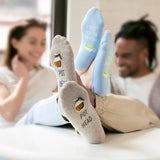 A person lying down with their feet raised, wearing gray socks printed with a beer mug and the words 'POT HEAD' and light blue socks printed with a beer mug and the words 'I LOVE YOU SO MATCHA'. Two blurred people are visible in the background. thumbnail