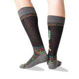 A pair of crew socks worn on a person's legs, featuring a dark charcoal gray base with a colorful geometric pattern in red, green, and teal, and a cactus motif on the side. thumbnail