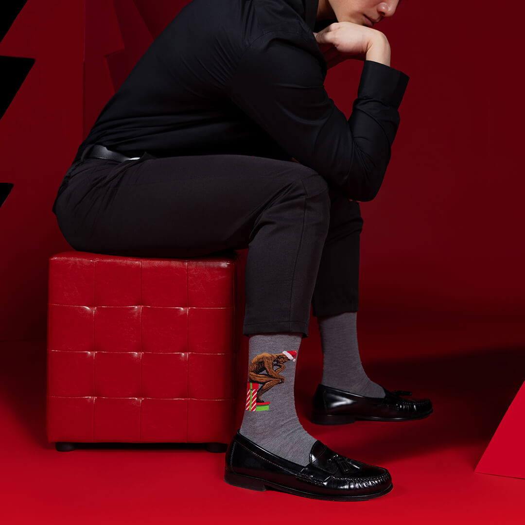 Person sitting on a red stool wearing black shoes and gray socks with The Thinker design, against a red background.