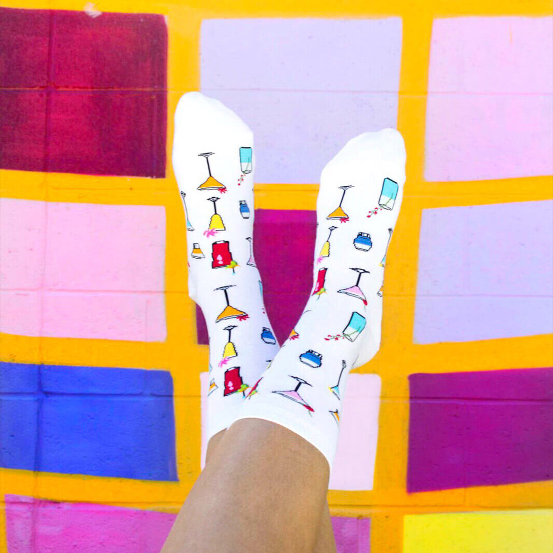 White crew socks with a colorful pattern of cocktail glasses, shown on a person's feet against a vibrant, multicolored tiled wall.