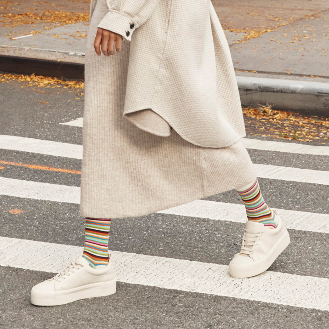 A person walking across a crosswalk wearing a long cream knit dress, a light beige cardigan, colorful striped socks, and white platform sneakers.