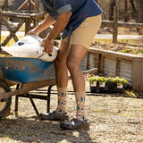 A person in a garden wearing beige crew socks with a colorful pine tree pattern, dark gray Crocs, and tan shorts, standing next to a blue wheelbarrow while holding a bag of soil or compost. thumbnail