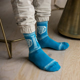 A person wearing bright blue crew socks with white Greek key and classical column patterns, standing on a light wood floor next to a brass chair.