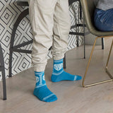 A person wearing bright blue crew socks with white graphic prints and beige jogger pants, standing on a light wood floor in a modern interior with patterned wallpaper and chairs.