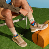 A person seated on a chair outdoors on artificial grass, wearing beige hiking socks with colorful mountain graphics and text, paired with tan and gray sneakers.