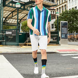 A man walking outdoors in front of a subway station wearing a short-sleeve polo shirt with vertical stripes in green, blue, white, and dark navy, white shorts, black socks with rainbow stripes, and white sneakers.