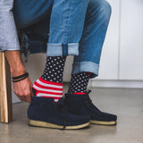 A person wearing blue jeans and dark navy suede boots, with one hand adjusting patriotic socks that feature a white star pattern on a black background and red-and-white stripes near the top. thumbnail