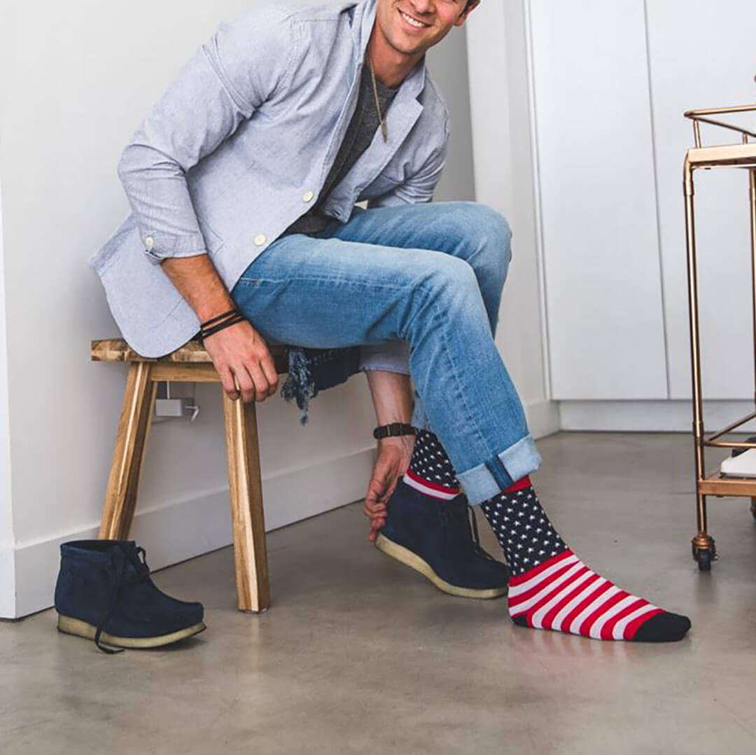 A man sitting on a wooden stool wearing blue jeans, a light blue shirt, and navy blue boots, while trying on a pair of socks with an American flag pattern. The socks feature red and white stripes on one leg and a navy blue background with white stars on the other, with red cuffs.
