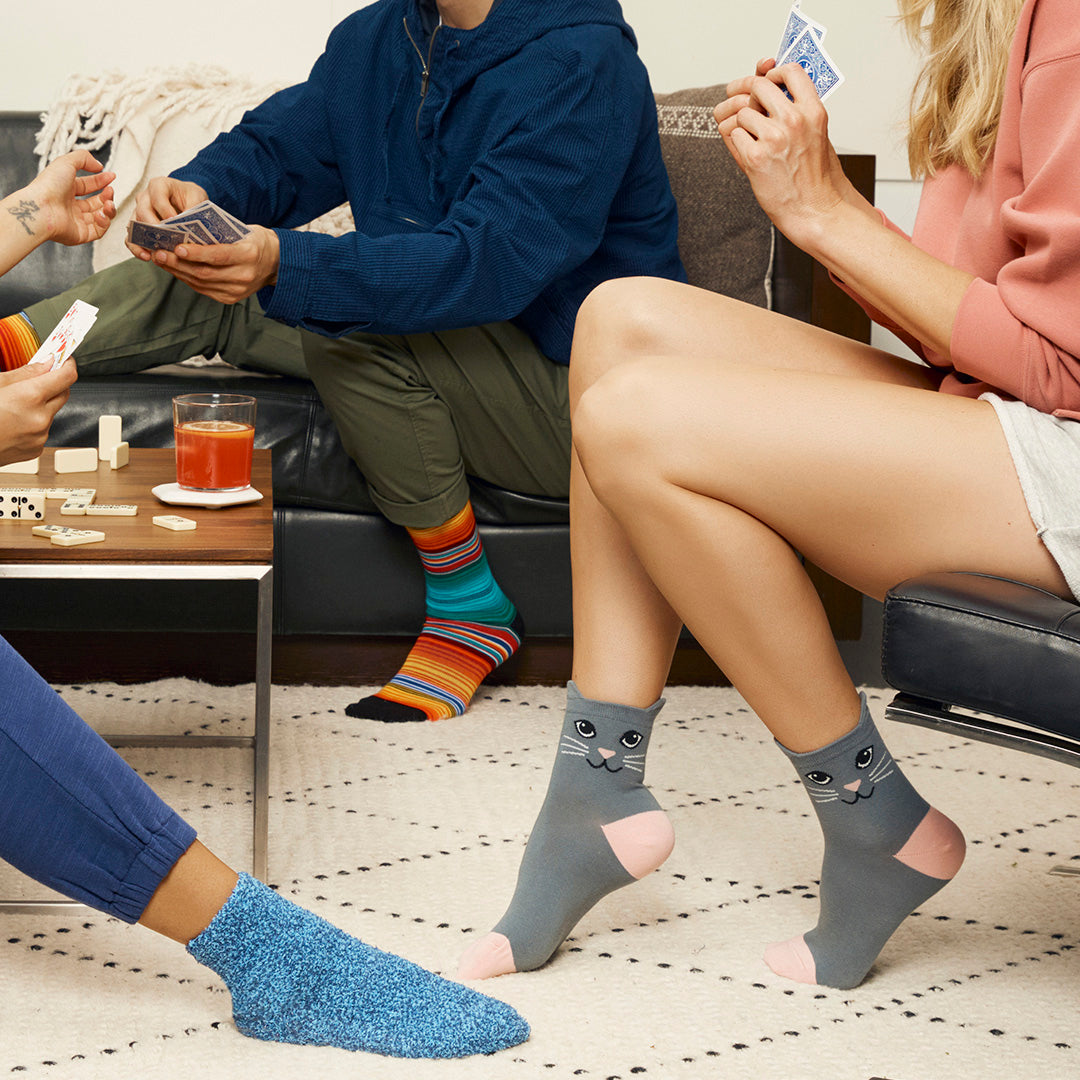 Gray ankle socks with a cat face design and pink toes, worn by a person sitting on a couch while playing cards in a cozy living room setting.