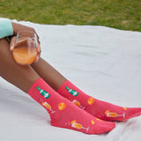 A person sitting outdoors on a white blanket holds a glass of orange drink while wearing bright pink crew socks printed with cocktail and citrus motifs. Grass is visible in the background. thumbnail