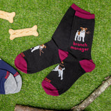 Black socks with dog illustrations and 'branch manager' text on a grass background with dog bones and a branch.