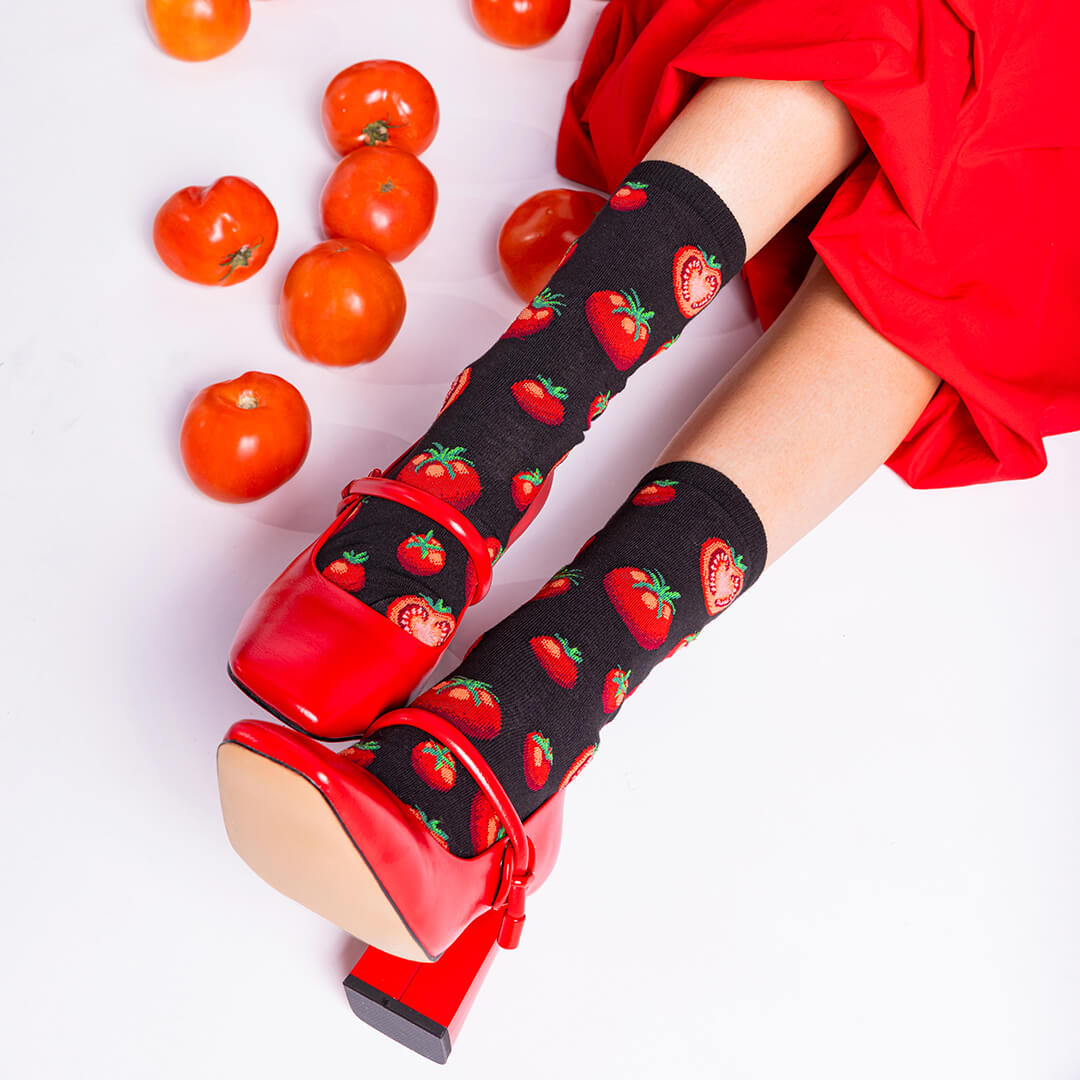 Person wearing black socks with red tomato pattern and red shoes, surrounded by tomatoes on a light background