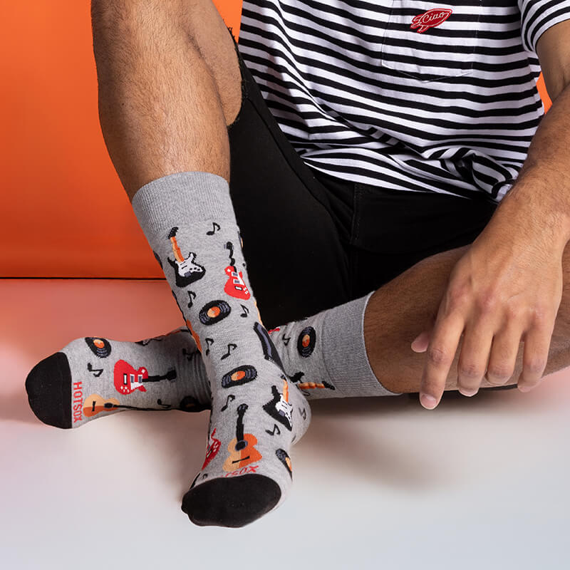 Person wearing gray socks with guitar and music note designs, sitting on a pink surface with an orange background.