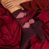A person wearing midi-crew argyle socks in shades of burgundy, dark red, and pink, resting on a draped burgundy satin fabric.