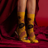 A pair of metallic gold crew socks with a dark floral pattern, worn with clear high-heeled shoes and shown on a model's feet against a draped dark red background. thumbnail