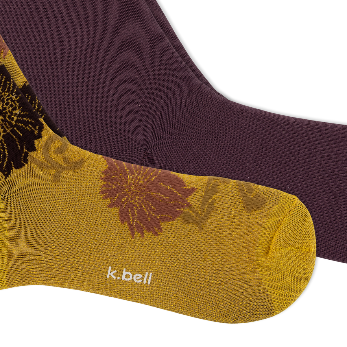 A close-up of two socks from k.bell, featuring a metallic gold sock with a floral pattern and white brand text, layered over a solid dark plum purple sock, on a white background.