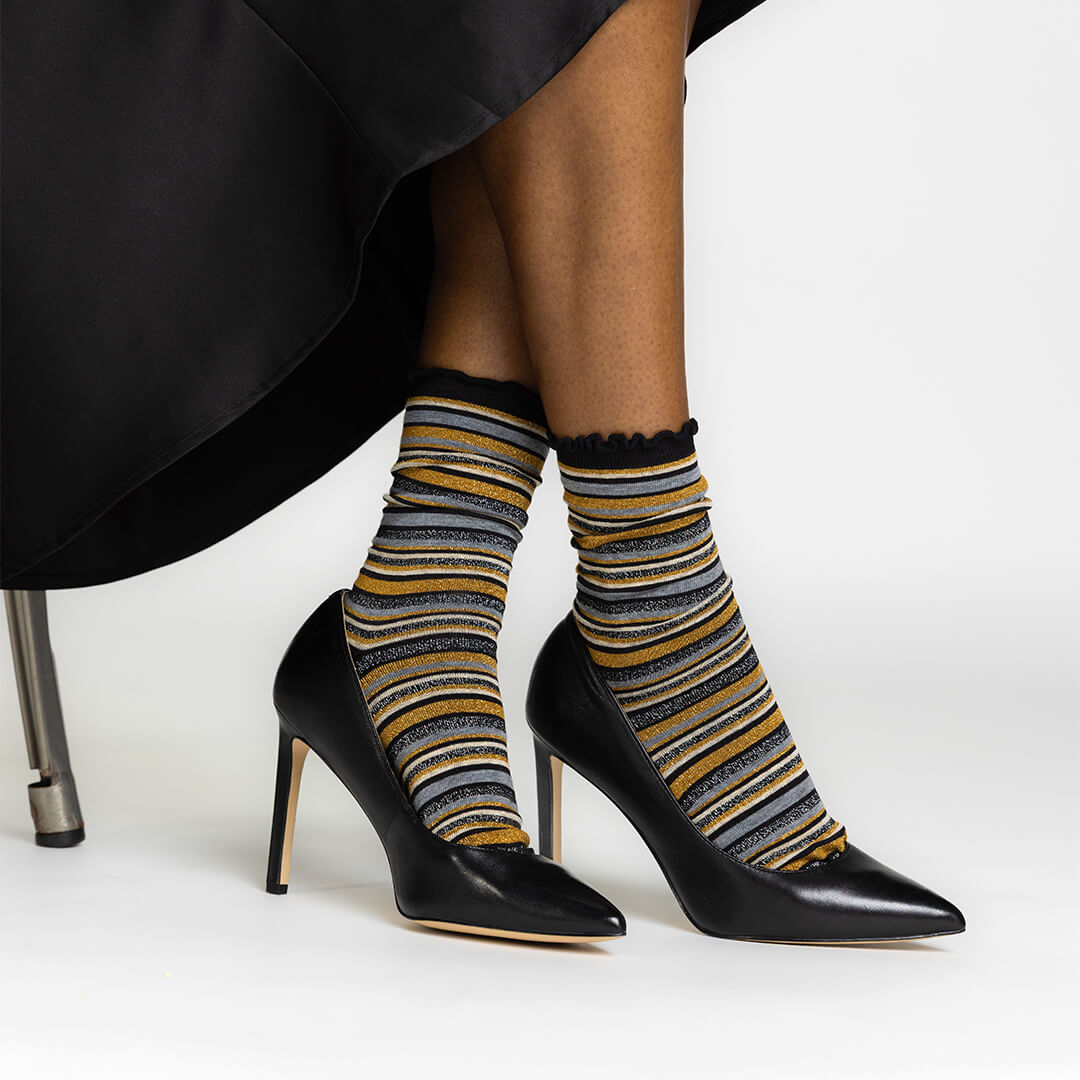 A person seated in a black chair wearing black pointed-toe high heels and metallic-striped crew socks with black ruffled tops. The socks have horizontal stripes in black, mustard yellow, gray, and white.
