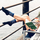 Person sitting by a waterfront holding a book, wearing blue patterned socks. thumbnail