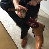 Person sitting on a chair wearing brown leather shoes and dark pants, holding a glass with a cigar.