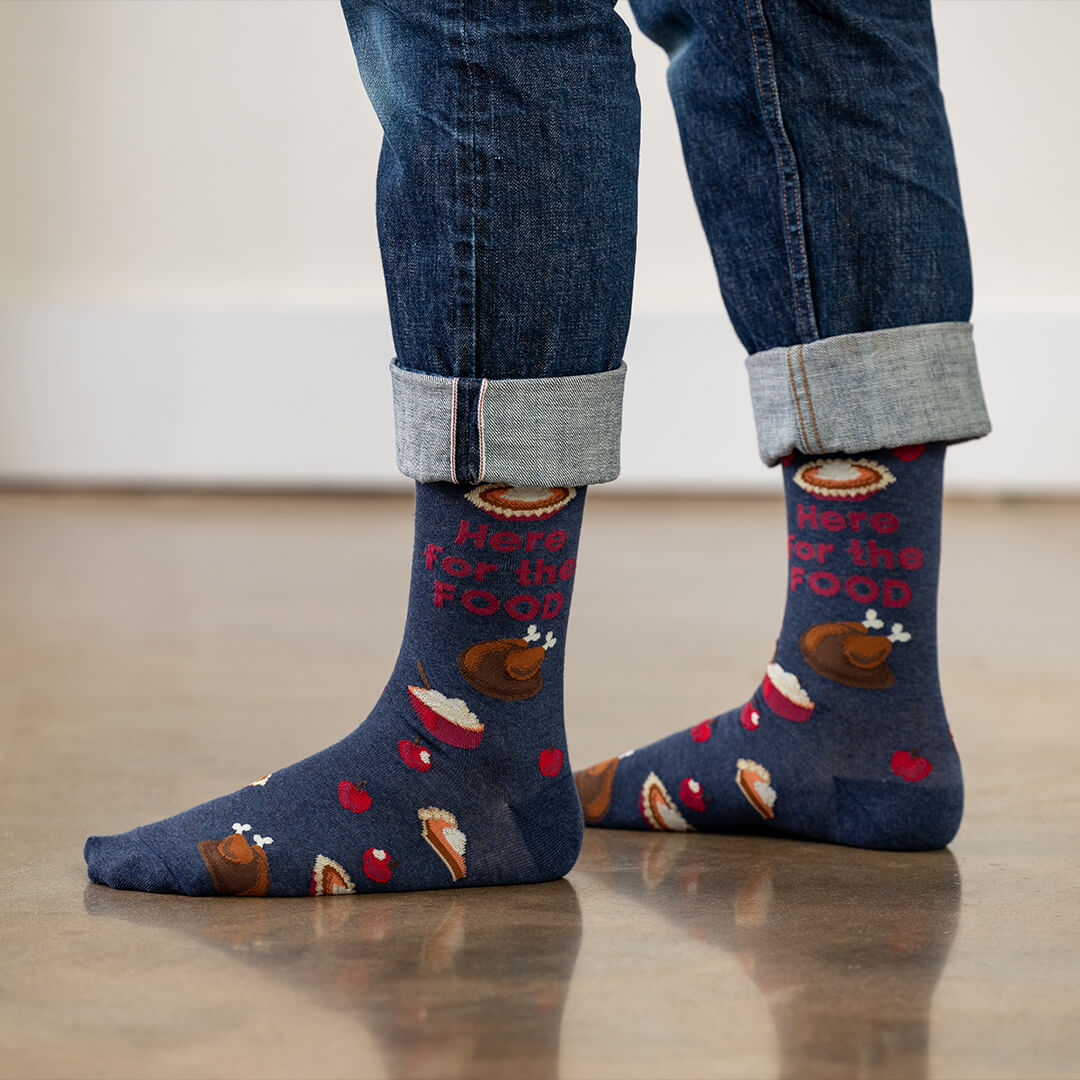 Person wearing blue socks with Thanksgiving-themed design and rolled-up jeans on a light wooden floor.