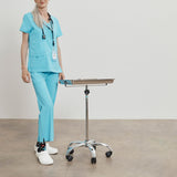 Person in light blue medical scrubs standing next to a height-adjustable table on a plain background thumbnail