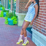 Woman in a light blue dress and yellow high-heeled shoes leaning against a brick wall with plants in the background.