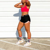 Person wearing a red sports bra and black shorts with 'Under Armour' branding, standing against a white wall.