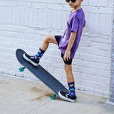 Child in purple shirt and black shorts standing on a skateboard against a white brick wall. thumbnail