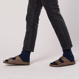 Person wearing brown sandals with blue socks and dark jeans on a light gray background