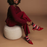 Woman in red outfit with striped socks and red high heels sitting on a white woven stool against a brown background thumbnail
