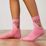 Pink socks with fist design and 'Women Unite' text worn by a person on a wooden floor.