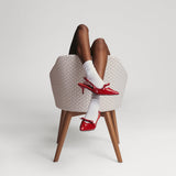 Person wearing red high-heeled shoes and white socks sitting on a woven stool against a plain background